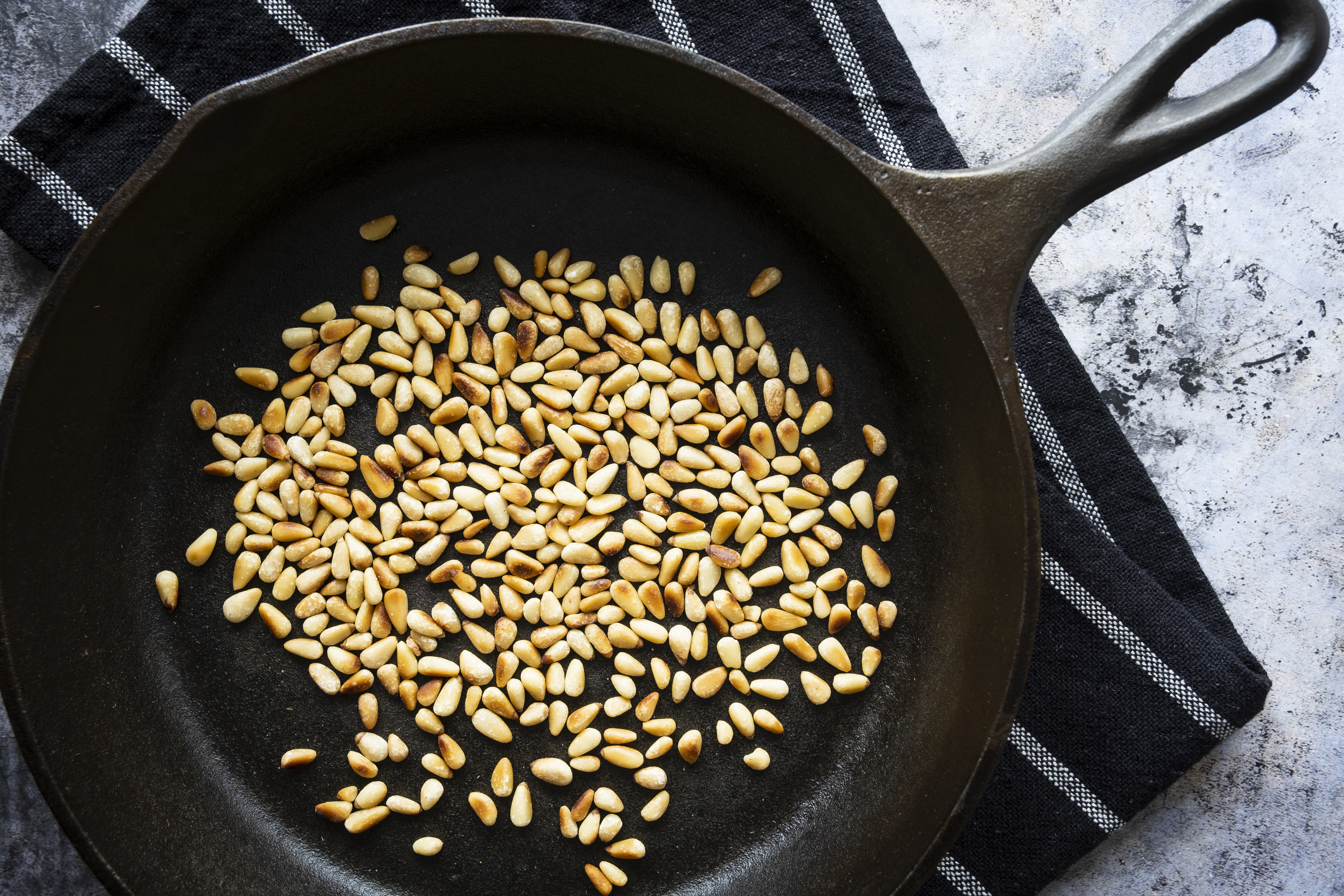 Toasted pine nuts in a cast iron skillet on a black tea towel.